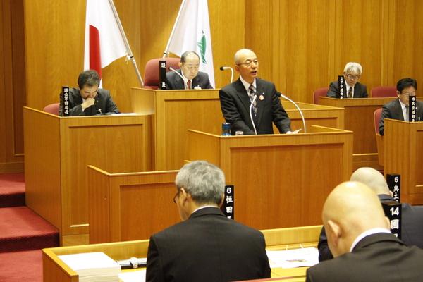 市長が議会にて答弁し、周りの議員が資料に目を通しながら熱心に聞き入っている様子の写真